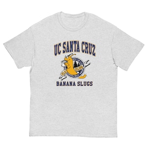 May include: A light gray short-sleeve t-shirt featuring a graphic of a yellow banana slug wearing glasses and holding a book. The text reads UC SANTA CRUZ, BANANA SLUGS, and UNIVERSITY OF CALIFORNIA SANTA CRUZ within a circular emblem design.