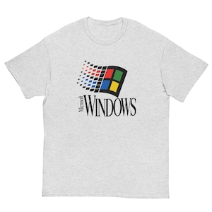 Puede incluir: Una camiseta gris claro de manga corta con el logotipo clásico de Microsoft Windows en el pecho. El gráfico muestra una ventana estilizada con cuadrados rojos, verdes, azules y amarillos, acompañada por el texto negro Microsoft WINDOWS.