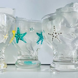 Könnte beinhalten: Vier klare Glasbecher mit bemalten Seestern-Designs. Die Becher haben eine breite Schale und einen Stielsockel. Die Seesterne sind in Gelb, Türkis, Grau und Rot bemalt.