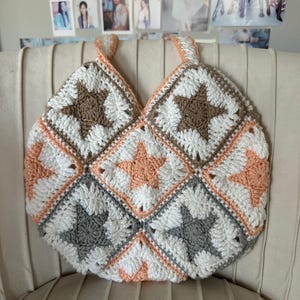 Peut inclure: Un sac fourre-tout crocheté à motifs d'étoiles en blanc, marron, gris et pêche. Le sac a deux anses et est posé sur une chaise beige. Le sac est fait de fil.