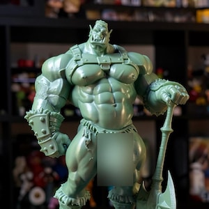 Könnte beinhalten: Grüne Figur eines muskulösen Ork-Kriegers, der eine Axt hält. Der Ork hat detaillierte Muskeln, Stoßzähne und spitze Ohren. Die Figur steht auf einem schwarzen, kreisförmigen Sockel, der ein Fantasy-Thema zeigt.