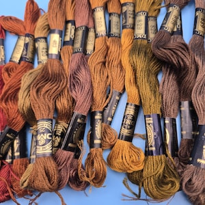 May include: A selection of DMC embroidery threads in shades of brown, orange, and green. The spools are labelled with numbers and brand details, set against a light blue backdrop. Suitable for cross-stitch and embroidery work.