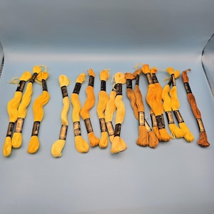 以下が含まれることがあります： 黄色とオレンジ色の刺繍糸の束のコレクション。 糸は波状に配置され、各束には白黒のタグが付いています。 色は、明るい黄色から深いゴールデンブラウンまであります。