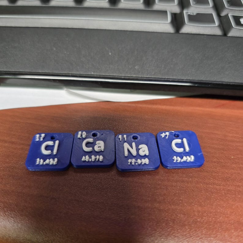 以下が含まれることがあります： 白文字と数字が入った青い正方形のペンダント4つ。ペンダントには、化学記号Cl、Ca、Na、Clと原子番号と重量が表示されています。それぞれ上部に小さな穴があります。