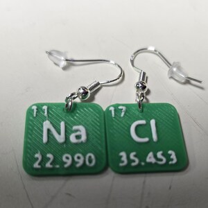 以下が含まれることがあります： 緑色の正方形のペンダントが付いた、銀色のイヤリング。各ペンダントには、白い化学記号と原子量が表示されています。Na（22.990）とCl（35.453）。イヤリングはポストとフックのデザインです。