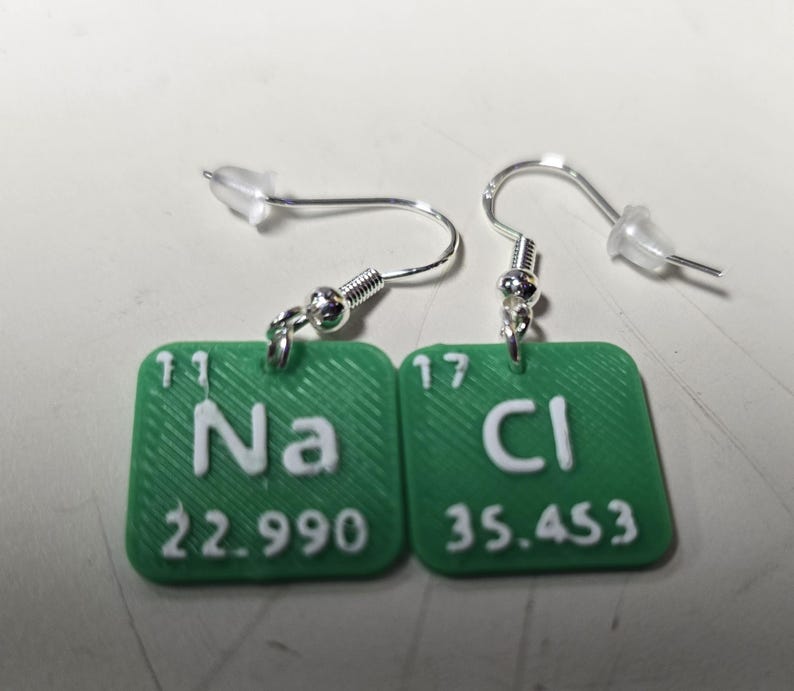 以下が含まれることがあります： 緑色の正方形のペンダントが付いた、銀色のイヤリング。各ペンダントには、白い化学記号と原子量が表示されています。Na（22.990）とCl（35.453）。イヤリングはポストとフックのデザインです。