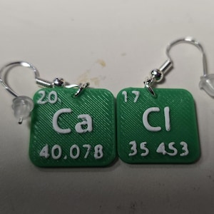以下が含まれることがあります： 周期表の化学記号「Ca」と「Cl」が描かれた緑色のイヤリングのペア。各イヤリングは正方形で、白い文字と銀色のフックが付いています。原子番号と重量も表示されています。