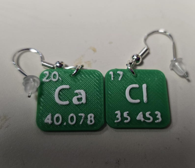 以下が含まれることがあります： 周期表の化学記号「Ca」と「Cl」が描かれた緑色のイヤリングのペア。各イヤリングは正方形で、白い文字と銀色のフックが付いています。原子番号と重量も表示されています。