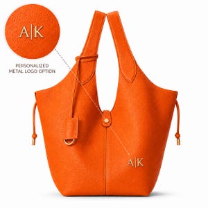 May include: Bright orange leather tote bag with a personalized metal logo option. The bag features a shoulder strap, a small tag with a metal ring, and drawstring details. The initials "AK" are displayed in gold.