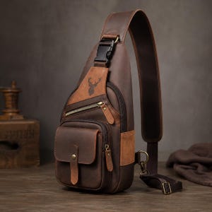 Pode incluir: Uma bolsa tiracolo de couro marrom com uma alça única e uma fivela de plástico preta. A bolsa possui um bolso frontal com fecho de pressão, um compartimento com zíper e um logotipo de cabeça de veado estampado na parte superior.