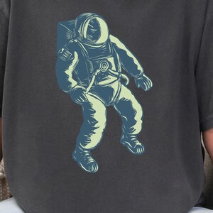 May include: Dark gray t-shirt featuring a blue and green astronaut graphic. The astronaut is wearing a spacesuit. The text "CC1717 PEPPER" is in the upper left corner. The shirt is a comfortable, casual style.
