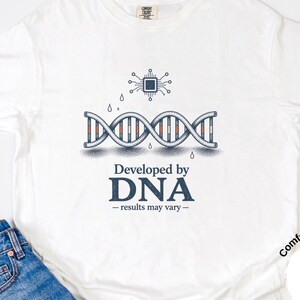 May include: White t-shirt with a graphic of a DNA strand and a circuit board, with the text "Developed by DNA - results may vary." The shirt is paired with blue jeans.
