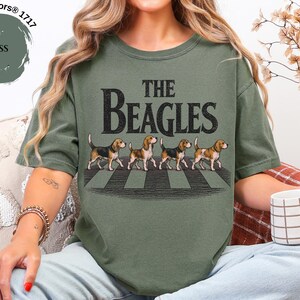 May include: Moss green t-shirt with the text "The Beagles" in a distressed font. The graphic features four beagle dogs walking across a crosswalk. The shirt is a Comfort Colors 1717 style.