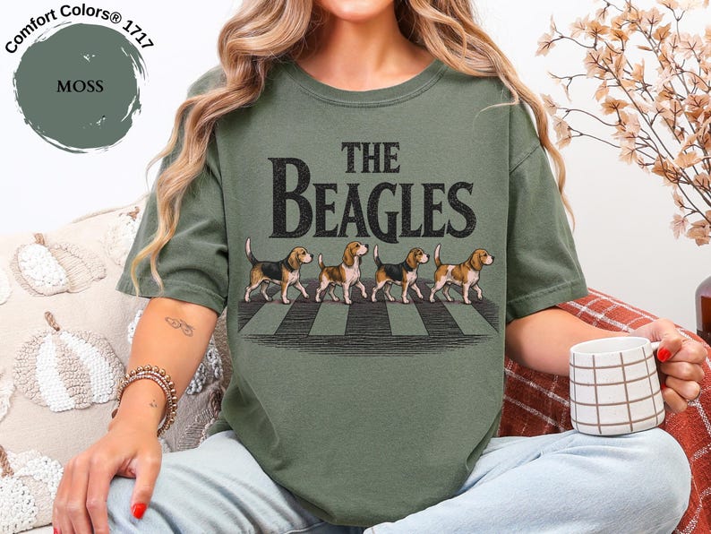 May include: Moss green t-shirt with the text "The Beagles" in a distressed font. The graphic features four beagle dogs walking across a crosswalk. The shirt is a Comfort Colors 1717 style.