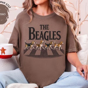 May include: Espresso-coloured t-shirt with the text "THE BEAGLES" and an illustration of four beagles crossing a crosswalk. A white mug with a red base and a gingerbread man design is held in the hand.