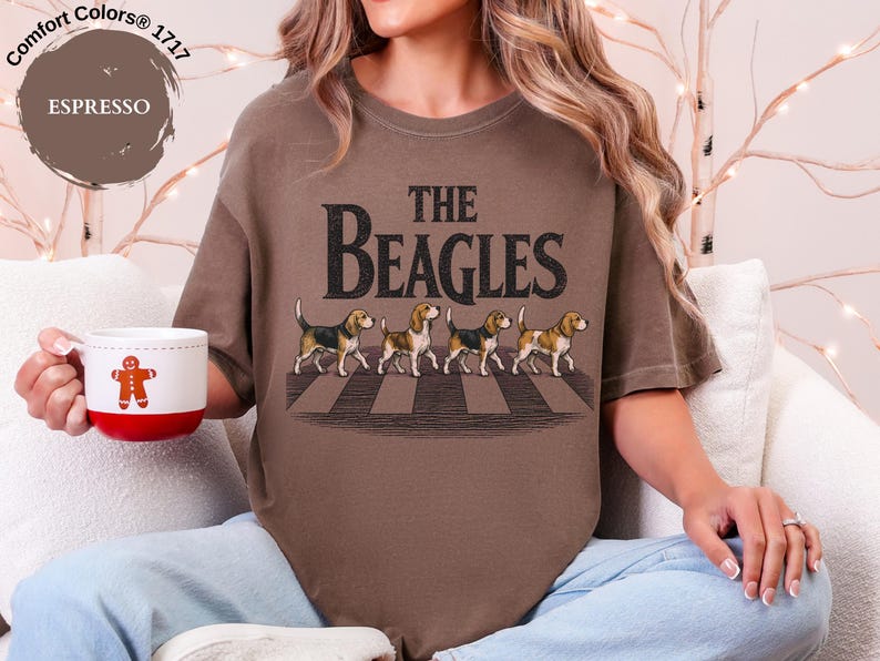 May include: Espresso-coloured t-shirt with the text "THE BEAGLES" and an illustration of four beagles crossing a crosswalk. A white mug with a red base and a gingerbread man design is held in the hand.