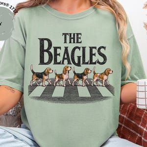 May include: A pale green t-shirt featuring the words "THE BEAGLES" and an image of four beagles crossing a zebra crossing. The shirt is a Comfort Colors 1717 style.