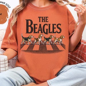 May include: A yam-coloured t-shirt with the text "THE BEAGLES" in black, featuring an illustration of four beagles crossing a crosswalk. The shirt is a Comfort Colors 1717 style.