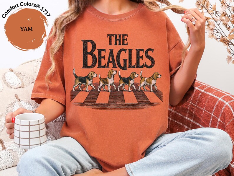 May include: A yam-coloured t-shirt with the text "THE BEAGLES" in black, featuring an illustration of four beagles crossing a crosswalk. The shirt is a Comfort Colors 1717 style.