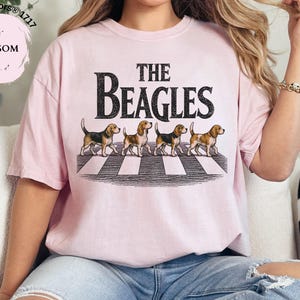 May include: A pale pink t-shirt featuring the words "THE BEAGLES" in black, with four beagle dogs on a zebra crossing. The shirt is a Comfort Colors 1717, Blossom.
