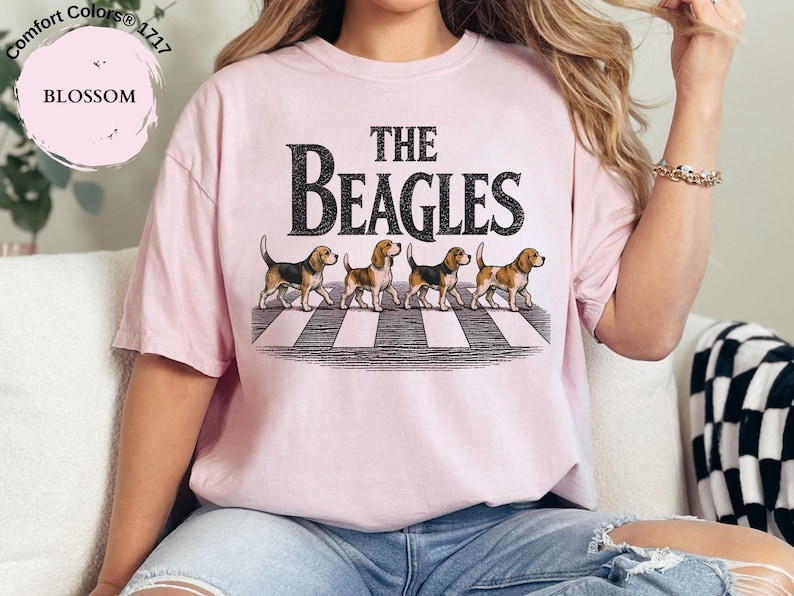 May include: A pale pink t-shirt featuring the words "THE BEAGLES" in black, with four beagle dogs on a zebra crossing. The shirt is a Comfort Colors 1717, Blossom.