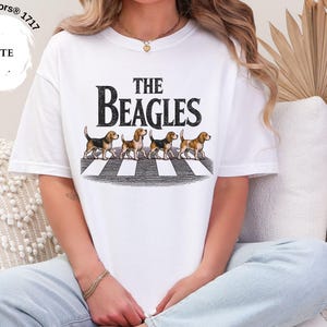 May include: White t-shirt with the words "THE BEAGLES" in a black font above an illustration of four beagles on a crosswalk. The shirt is a Comfort Colors 1717 style.