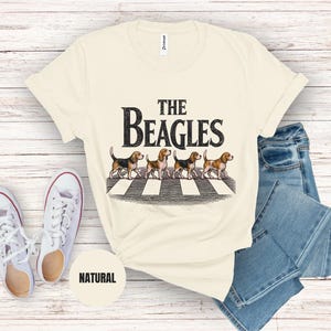 May include: A cream-coloured t-shirt featuring the words "The Beagles" and an illustration of four beagles on a zebra crossing. The shirt is styled with blue jeans and white trainers.