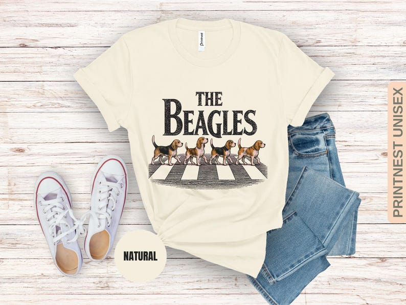 May include: A cream-coloured t-shirt featuring the words "The Beagles" and an illustration of four beagles on a zebra crossing. The shirt is styled with blue jeans and white trainers.