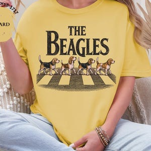 May include: Mustard yellow t-shirt featuring the text "THE BEAGLES" above an illustration of four beagles on a zebra crossing. The shirt is a Comfort Colors 1717 style.