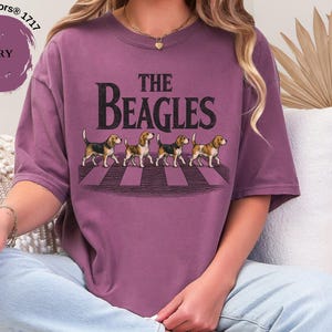 May include: Berry-coloured t-shirt with the words "The Beagles" and an image of four beagles on a zebra crossing. The shirt is a Comfort Colors brand.