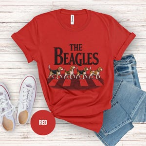 May include: Red t-shirt featuring the text "THE BEAGLES" and a graphic of four beagles on a crosswalk. The image includes blue jeans and white trainers. A red circle with the word "RED" is also visible.