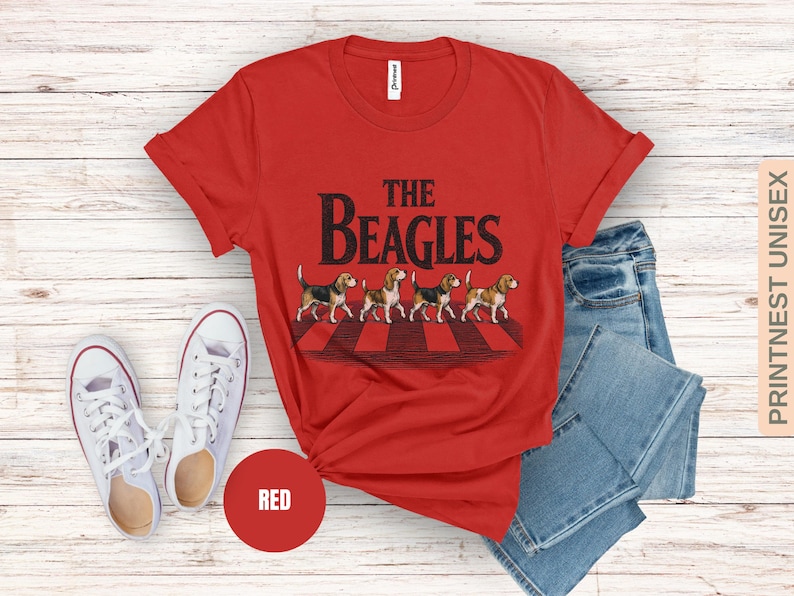 May include: Red t-shirt featuring the text "THE BEAGLES" and a graphic of four beagles on a crosswalk. The image includes blue jeans and white trainers. A red circle with the word "RED" is also visible.
