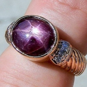 May include: A gold ring with a large, round, dark pink star ruby gemstone. The ruby has a star-like pattern in the center. The ring has a twisted band with small diamonds on the sides.