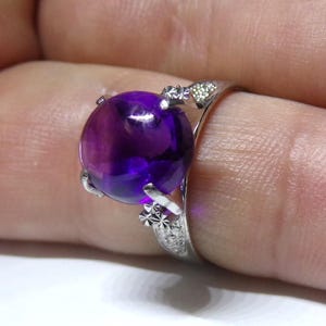 Puede incluir: Un anillo de plata con una gran amatista redonda de color p&uacute;rpura. El anillo tiene intrincados detalles de plata alrededor de la piedra.