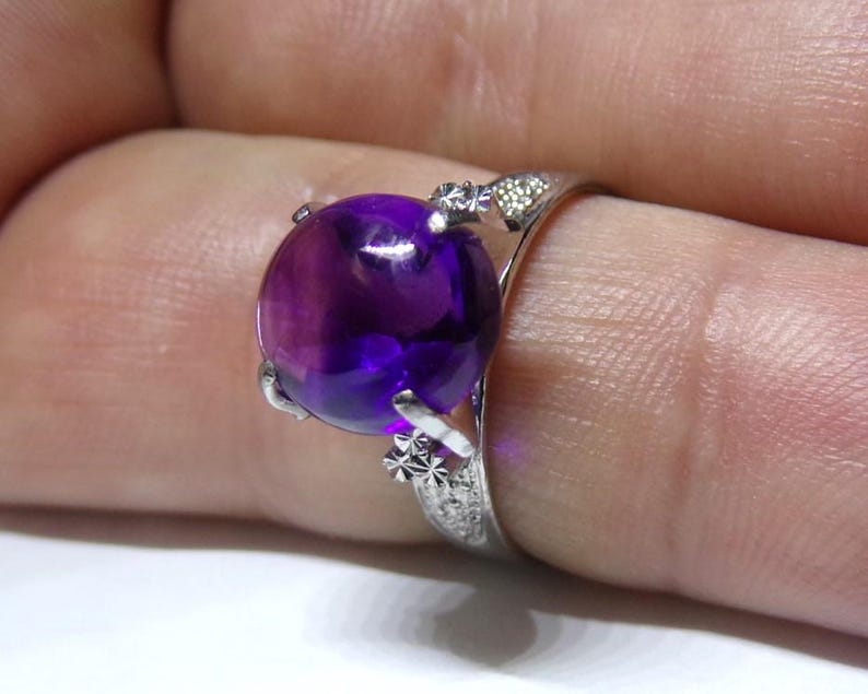 Puede incluir: Un anillo de plata con una gran amatista redonda de color p&uacute;rpura. El anillo tiene intrincados detalles de plata alrededor de la piedra.
