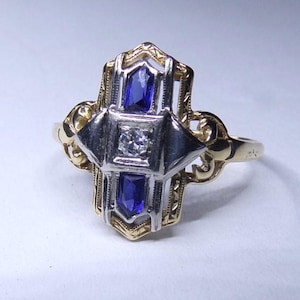 May include: A vintage ring with a geometric design featuring a central diamond and two blue sapphires. The ring is made of yellow and white gold.