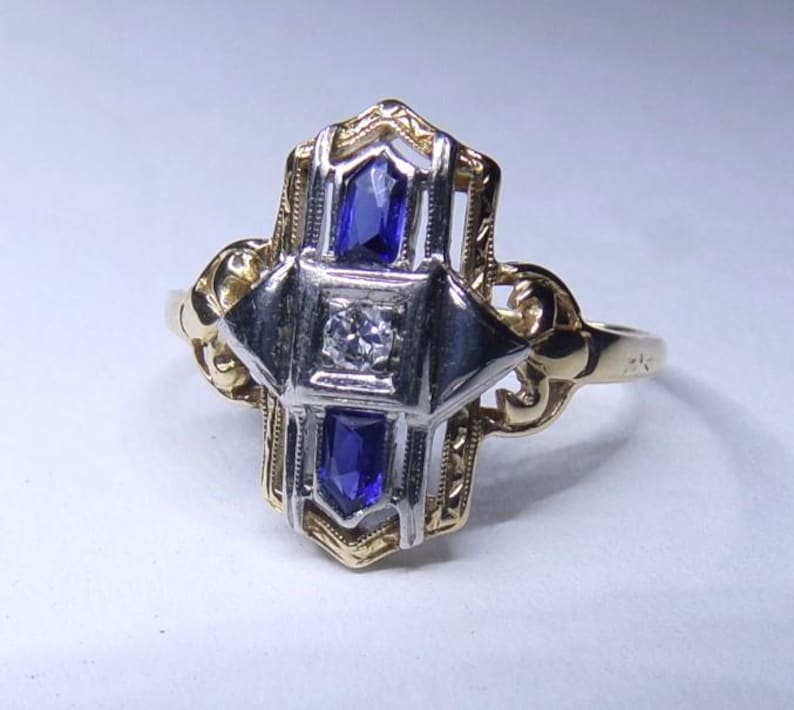 May include: A vintage ring with a geometric design featuring a central diamond and two blue sapphires. The ring is made of yellow and white gold.