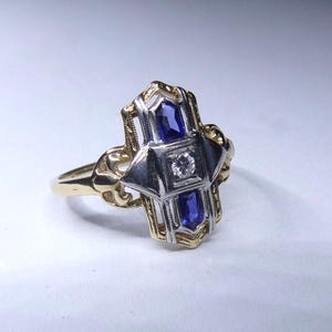 May include: A vintage ring with a geometric design featuring two blue sapphires and a single diamond in the center. The ring is made of yellow and white gold.