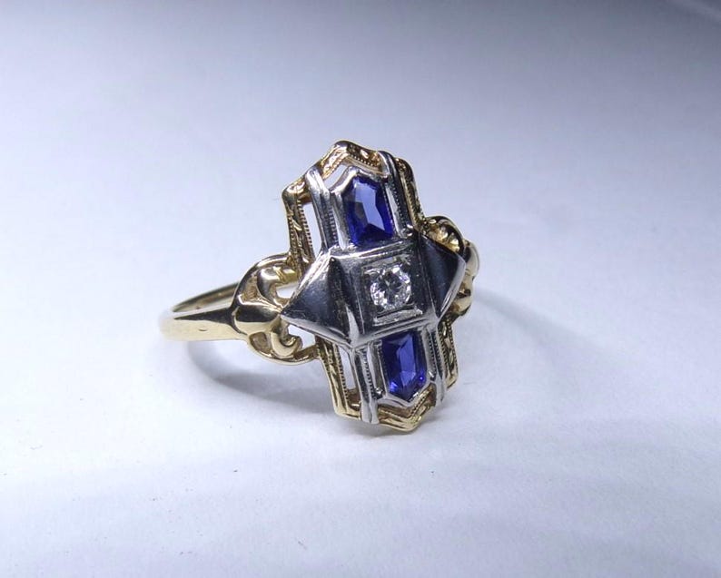 May include: A vintage ring with a geometric design featuring two blue sapphires and a single diamond in the center. The ring is made of yellow and white gold.