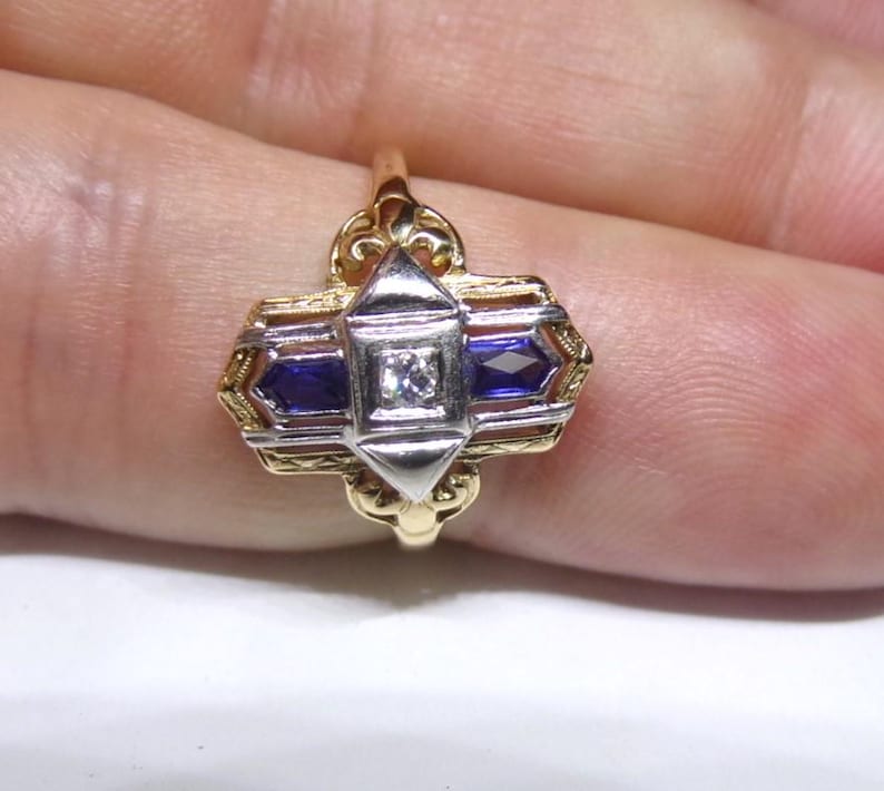 May include: A gold and silver ring with a geometric design. The ring features two blue sapphires and a single diamond in the center.