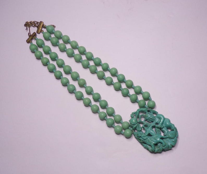 May include: A double strand necklace made of light green beads with a carved green pendant featuring a stylized figure.