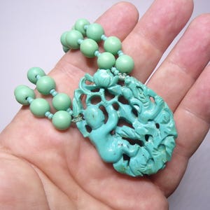 May include: A turquoise green necklace with a carved pendant featuring a floral design and two figures. The necklace is made of round beads.