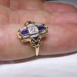 May include: A gold and silver ring with a geometric design featuring a central diamond and two blue sapphires.