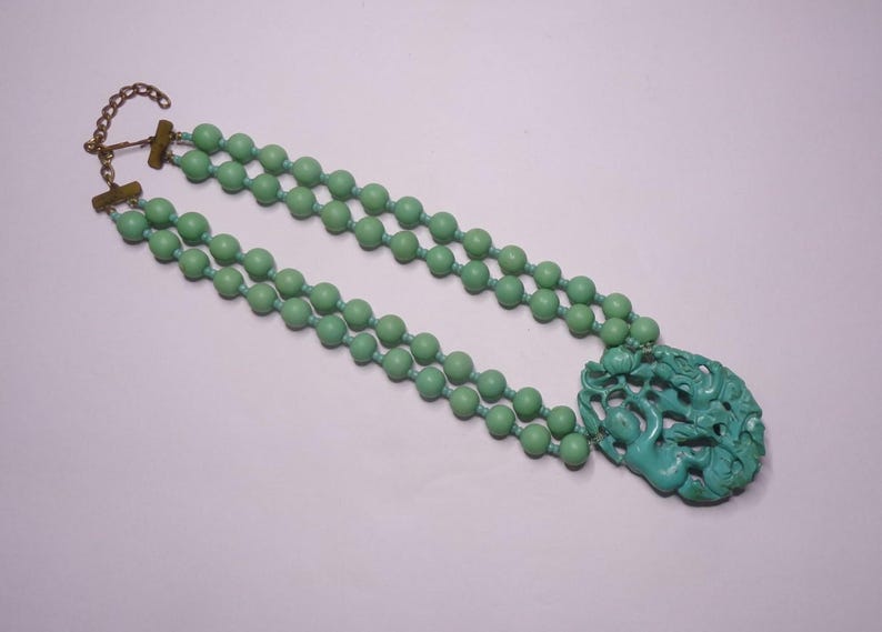 May include: A double strand necklace with a carved jade pendant. The necklace is made of light green beads and has a gold clasp.