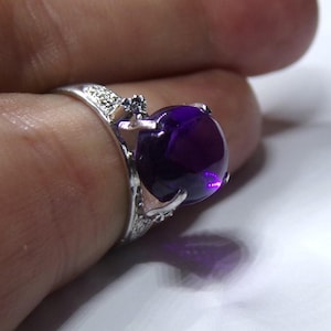 Puede incluir: Un anillo de plata con una gran amatista redonda de color p&uacute;rpura. El anillo tiene un dise&ntilde;o delicado y adornado con peque&ntilde;as piedras transparentes.