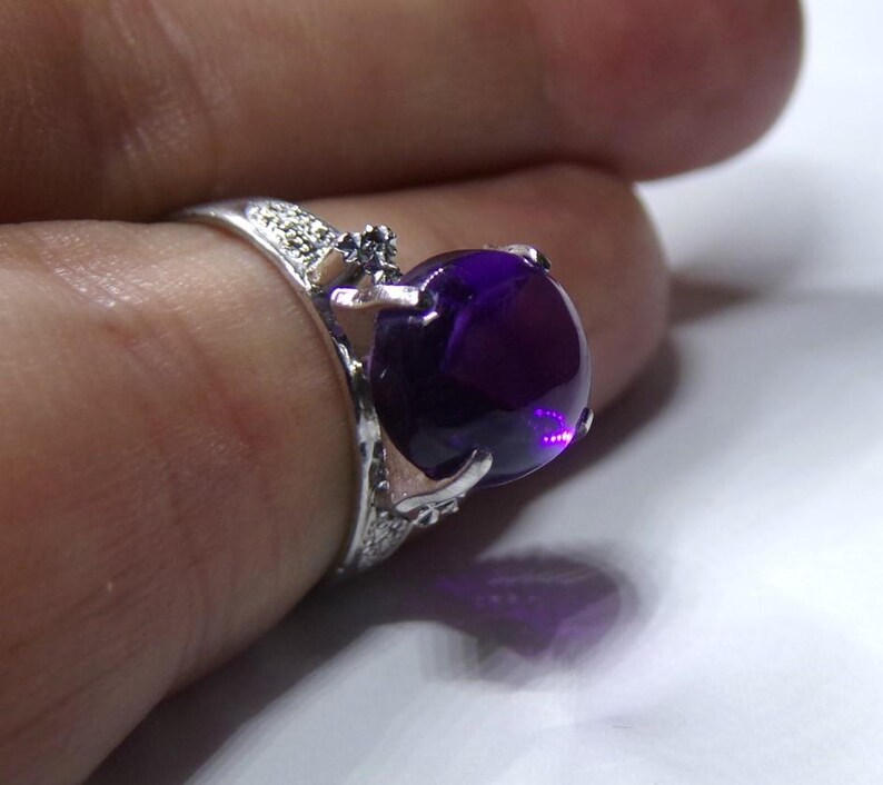 Puede incluir: Un anillo de plata con una gran amatista redonda de color p&uacute;rpura. El anillo tiene un dise&ntilde;o delicado y adornado con peque&ntilde;as piedras transparentes.