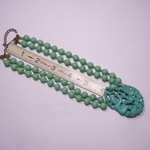May include: A turquoise green beaded necklace with a carved turquoise green pendant. The necklace is made of three strands of beads and the pendant is a detailed carving of a mythical creature.