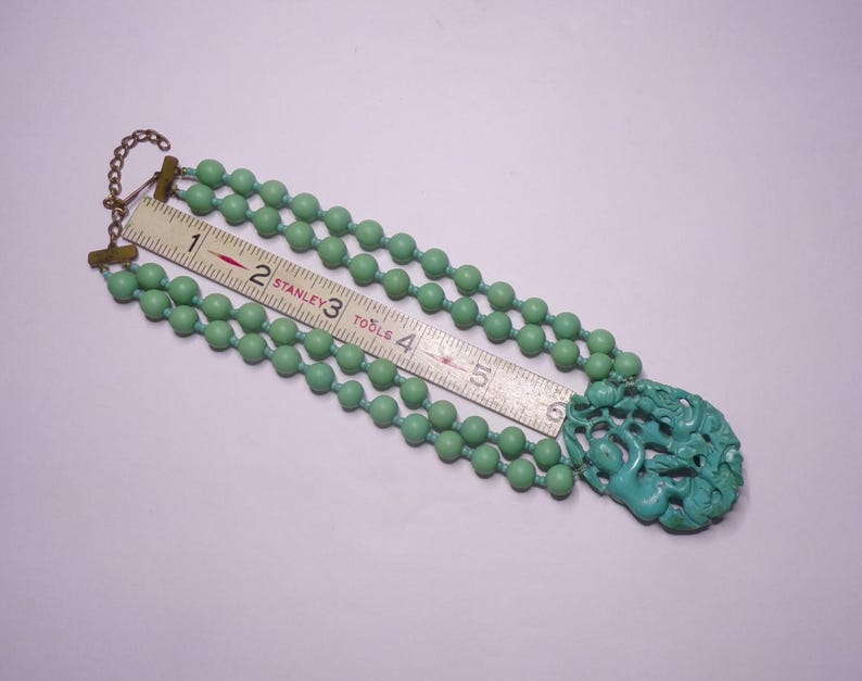 May include: A turquoise green beaded necklace with a carved turquoise green pendant. The necklace is made of three strands of beads and the pendant is a detailed carving of a mythical creature.