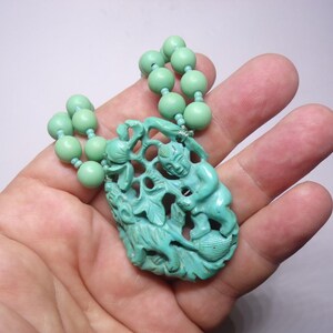 May include: A turquoise green necklace with a carved pendant featuring two figures and a leafy design. The necklace is made of round beads.