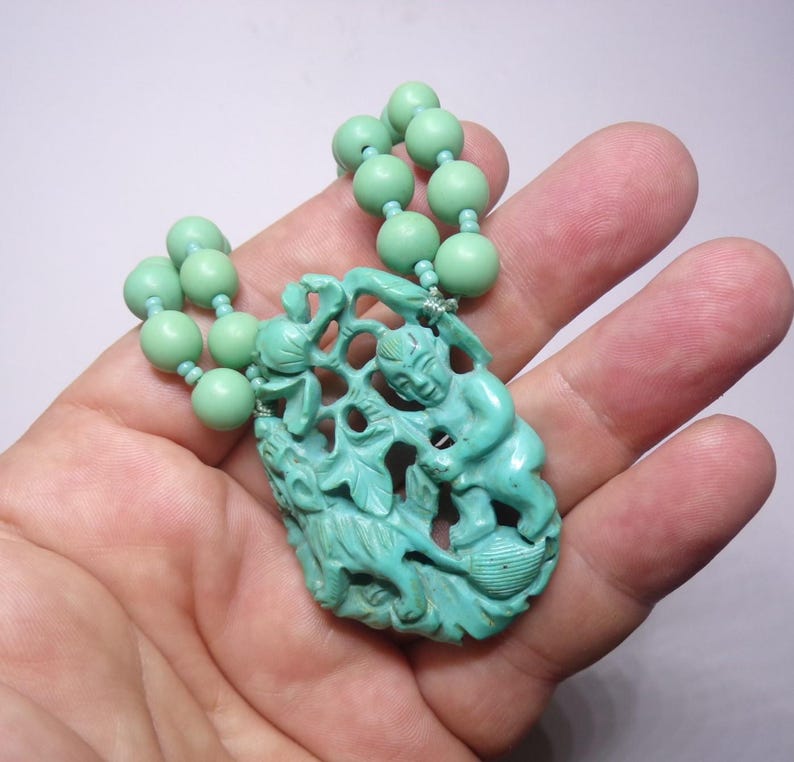 May include: A turquoise green necklace with a carved pendant featuring two figures and a leafy design. The necklace is made of round beads.